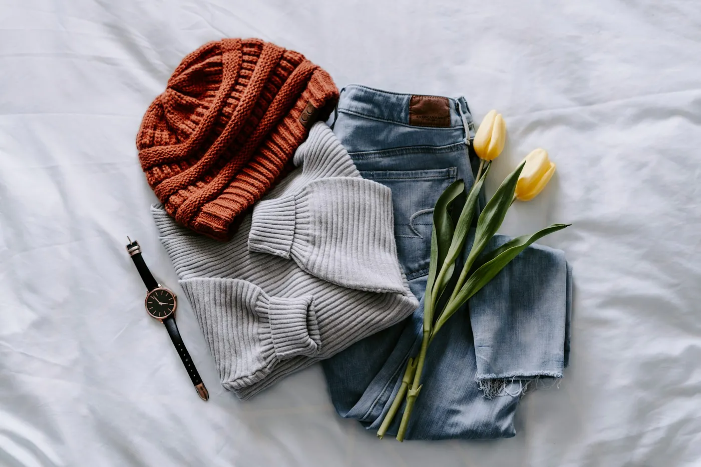 A flat lay of a knitted hat, jumper, jeans, a watch and tulips on white linen