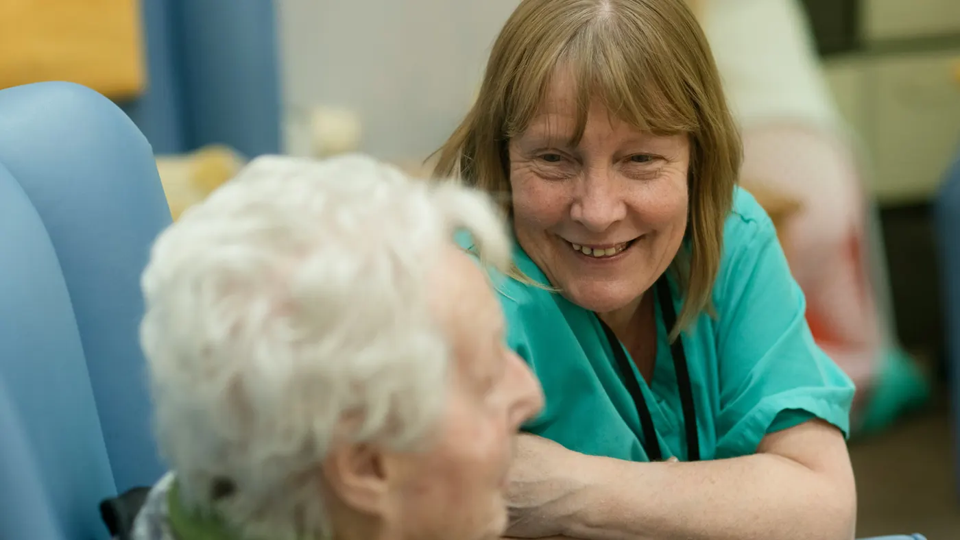 A smiling nurse in green scrubs leaning in to talk with an older woman