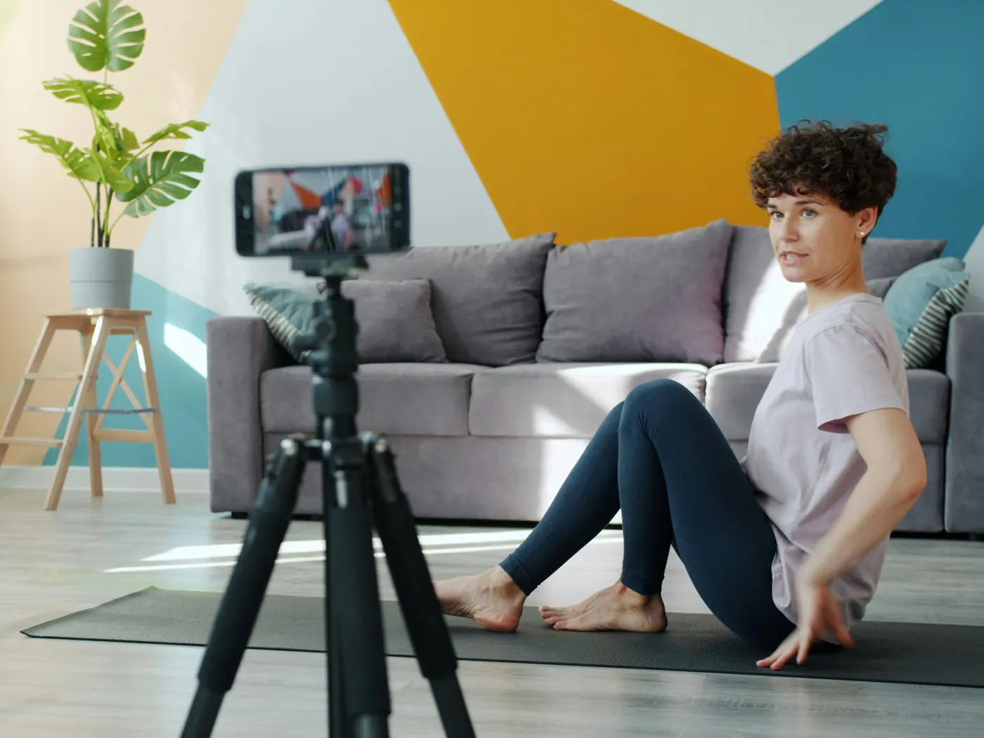 A woman on an exercise mat at home, with a phone on a tripod recording her movements