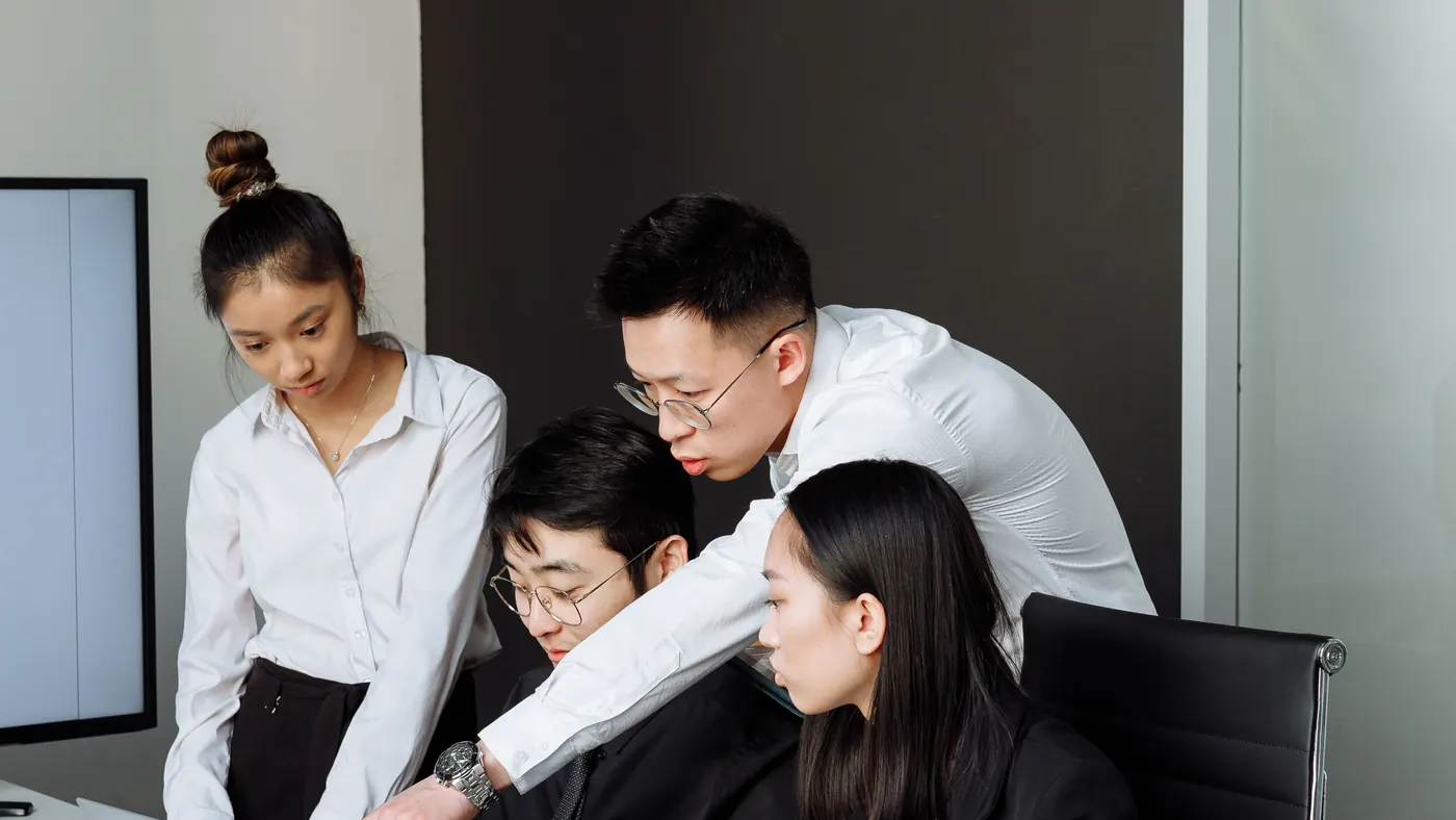 Four colleagues gathered around a laptop in a meeting room, reviewing something together