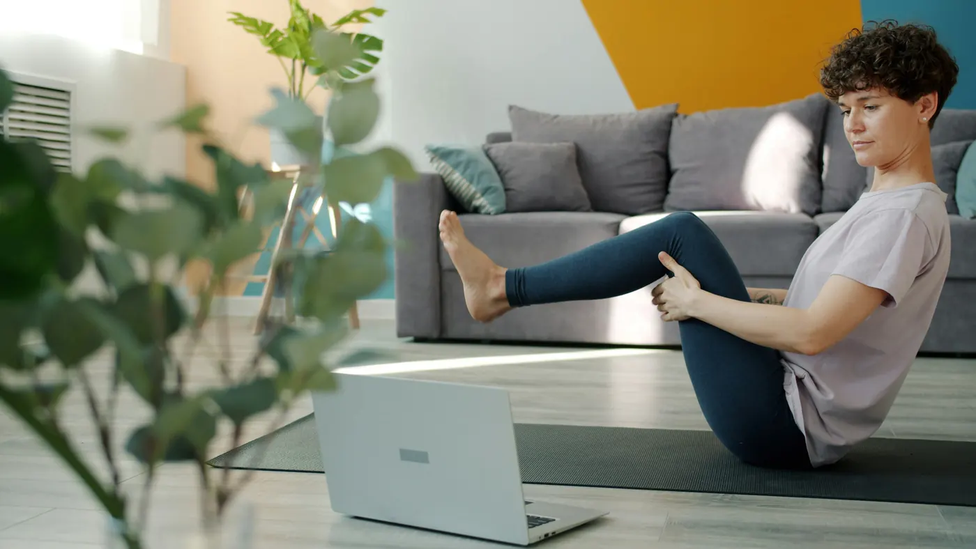 A woman doing a leg exercise on a mat at home, with a laptop open in front of her