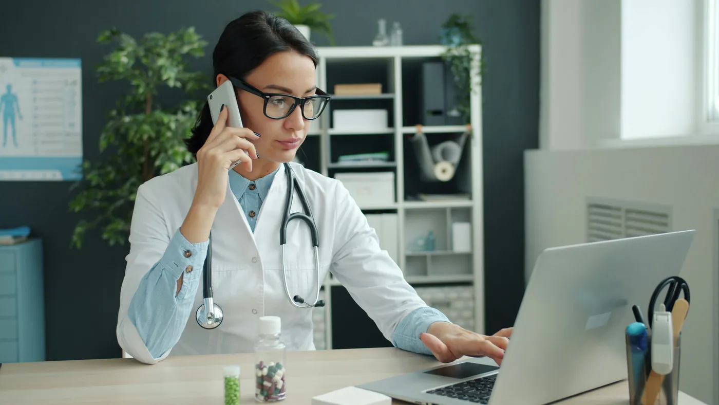 A doctor at her desk on the phone while typing on a laptop